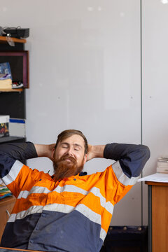 Man sitting behind the desk in high visibility clothes with eyes closed and hands behind his head.