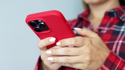 Young woman in a plaid shirt holding a red mobile phone in her hands typing a message on a grey background. A girl chatting with friends