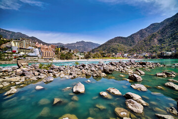 Scenic view of the Ganges River in Rishikesh, India, with clear turquoise waters, rocky shores, lush green mountains, and iconic temples in the background.