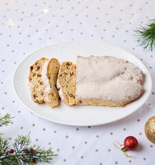 Baked Stollen or German Christmas cake, sprinkled with powdered sugar and sliced, on a white oval plate with a tablecloth in the background. Advent. Merry Christmas.