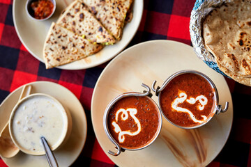 Overhead shot of traditional Indian meal with Dal Makhani, yogurt dip, naan, and papad on a plate, served on a red checkered tablecloth for a cozy dining experience.