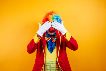 Playful clown hides face, bright yellow background.