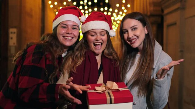 Three joyful young women celebrating Christmas, wearing festive outfits and Santa hats, smiling and holding a wrapped gift, enjoying the holiday spirit together under glowing decorative lights