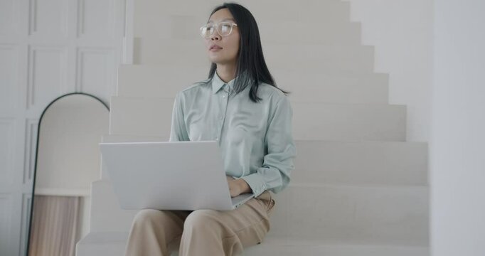 Beautiful Asian lady working with laptop typing sitting on stairs in stylish luxury house. Computer and elegant interior concept.