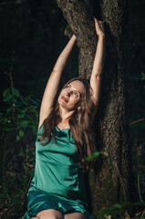 Dreamy smiling woman resting near tree with rough bark, looking up and hugging trunk. Green dress, long hair.