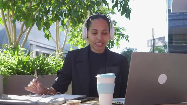 young latin business woman on a video call, sitting in an outdoor restaurant with her laptop, drinking coffee and taking notes.