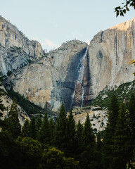 Waterfall cascading down a rugged cliff surrounded by pine trees. Mountain landscape under a clear...