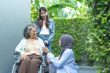 Two women, one in a hijab, supporting and interacting with an elderly woman in a wheelchair...