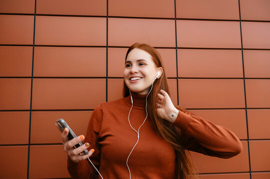Happy beautiful redhead woman listening to music in front of brown wall
