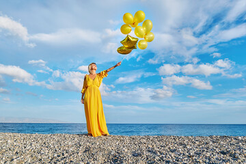 Mature woman in yellow dress standing with balloons at beach under cloudy sky