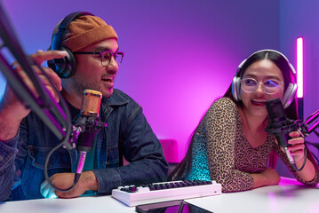 Two young asian streamer host making live podcast with condenser microphone and tablet at broadcast home studio. Content creator and social media influencer creating digital content.