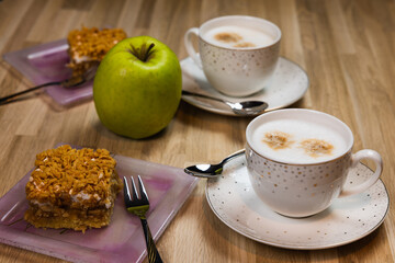 Two cups of cappuccino with spoons and an apple pie on plates ready to eat. A green apple lies next to it.