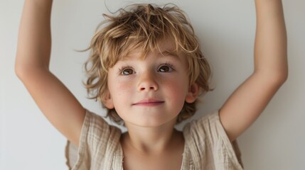 Naklejka premium A young boy with blonde hair and a beige shirt looking up with his arms raised.