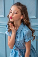 Fototapeta premium Young Girl in Denim Dress Blowing a playful Kiss on the blue paneled background.