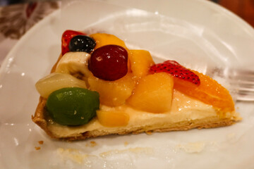 close up of a slice of mixed fruit cake tart with custard, grapes, mango, peach and cream with a fork on the plate