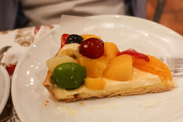 close up of a slice of mixed fruit cake tart with custard, grapes, mango, peach and cream with a fork on the plate