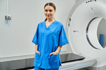 Confident female doctor in blue scrubs stands by an MRI machine, ready for diagnostics.