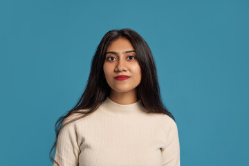 Portrait of 23 years old Indian woman with black hair, wearing light blouse, posing with calm face against blue studio background. Concept of human emotions, facial expression, youth