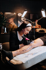 Charming girl tattoo on a man's leg in a tattoo parlor, creative small business and tattoo master. Hipster woman in gloves draws a tattoo