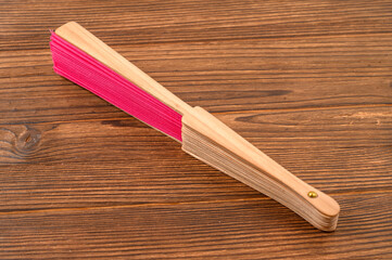 This beautifully crafted hand fan with a vibrant pink fabric, is folded and resting on a rustic wooden surface