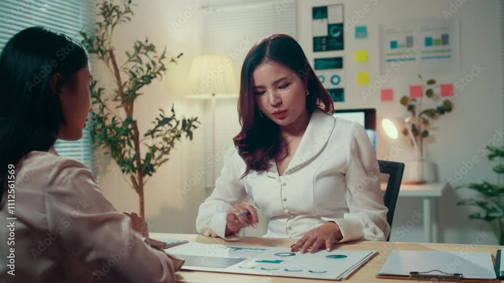 Aufkleber two businesswomen discuss financial charts at a desk in an office at night, with one using a calcula