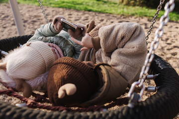 Fototapeta premium Cute little girls, two preschooler female kids uses the phone and headphone in the school yard. Concept image for child development, healthy kid playing outside. Gen Alpha. Part of a series
