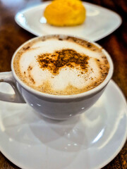 Close up, em xícara de louça branca com cappuccino e um pão de queijo ao fundo. Sobre a mesa de madeira.