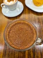 Close up no chocolate quente, cremoso, dentro de uma caneca de vidro, com uma xícara de louça branca com cappuccino ao fundo, sobre a mesa de madeira, em uma cafeteria.
