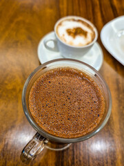 Close up no chocolate quente, cremoso, dentro de uma caneca de vidro, com uma xícara de louça branca com cappuccino ao fundo, sobre a mesa de madeira, em uma cafeteria.