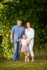 Fototapeta premium Joyful family posing happily holding hands in arms of greenery. Mom, dad, daughter relaxing together in park, having fun together. Vertical