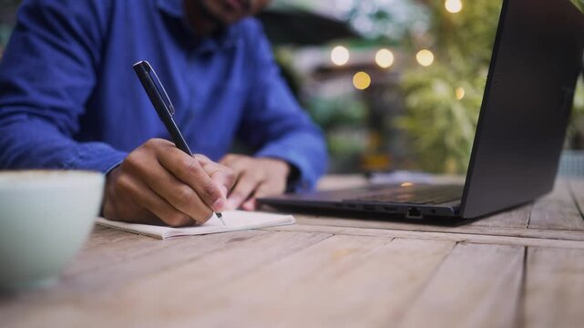 Close-up of a focused student or freelancer writing notes while working on a laptop outdoors. Unrecognizable individual in a casual setting taking notes by hand in a peaceful workspace.