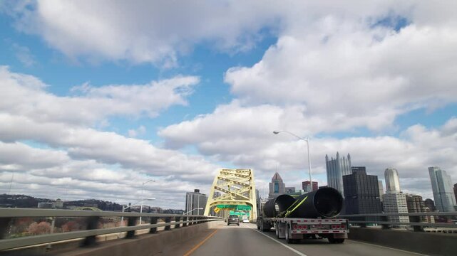 Car driver pov point of view moving toward Pittsburgh, PA city skyline and skyscraper buildings on multilane highway and interstate through vehicle traffic on golden bridge arches with blue sky