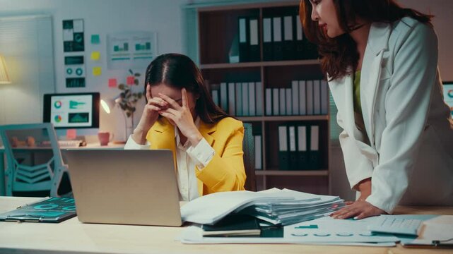Business woman is being reprimanded by her female manager. The employee is sitting at her desk with her head in her hands, while the manager stands over her, yelling