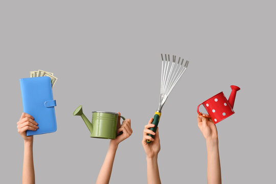 Female hands with watering cans, gardening rake and wallet on grey background