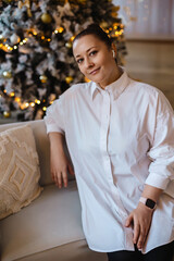 A confident woman in a loose white shirt sits on a cozy couch near a decorated Christmas tree with glowing lights. A festive and warm atmosphere perfect for holiday-themed visuals