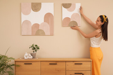 Young beautiful woman hanging picture on beige wall near chest of drawers in room