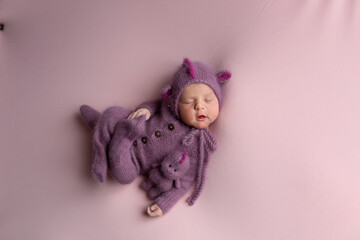Little adorable newborn baby girl sleeping on her stomach wearing a hat with dragon ears. Neutral background. Studio shot of a ten-day-old newborn baby. Beginning of life. Fairy tale. Dragon toy