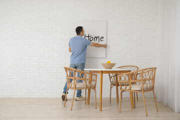 Young man hanging poster on white brick wall in dining room, back view