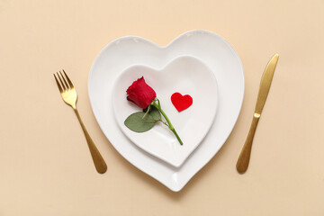 Table setting for Valentine's Day with heart-shaped plates, rose and paper heart on beige background