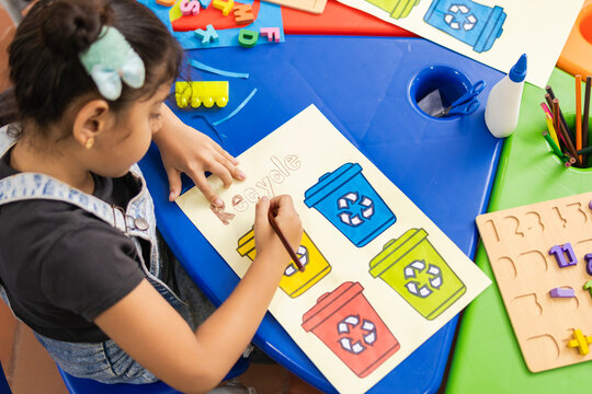 Latin girl in a primary school classroom, writing and painting the word "Recycle." Waste separation bins are visible as she learns about recycling