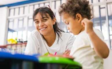 Young Latina teacher smiling at the camera while assisting a preschool boy with his homework in the classroom