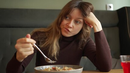 Young woman sits alone at a table, slowly eating her meal. Boredom and unhappiness are evident on her face as she engages in a solitary dining experience
