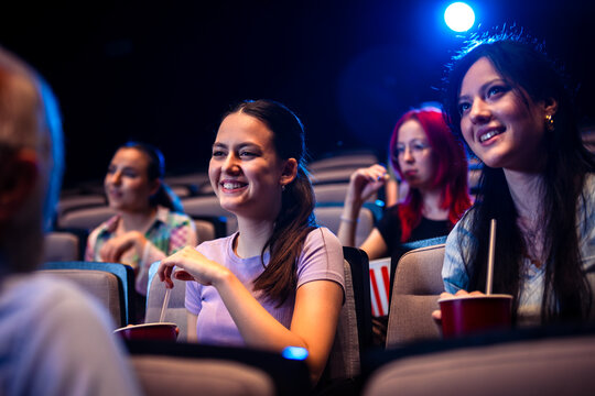 Two best friends are watching a movie in cinema and they are eating popcorn.