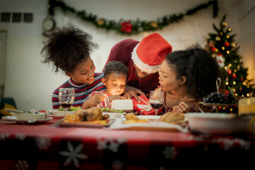 A joyful family gathers around the dinner table celebrating Christmas. The father, dressed in a Santa hat, gives a gift to the child, while everyone smiles with warmth and festive decorations.