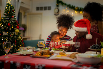 A joyful family gathers around the dinner table celebrating Christmas. The father, dressed in a Santa hat, gives a gift to the child, while everyone smiles with warmth and festive decorations.