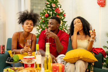 A group of friends celebrates Christmas together at home, enjoying drinks and laughter around a cozy living room. A decorated Christmas tree lights up the background, adding warmth to the holiday.