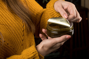 Hands in a yellow knitted sweater are peeling back the foil seal of a jar containing green cream. The image focuses on the product's packaging and the act of opening it.