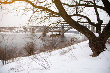 Rustic wooden bird feeder hanging from a tree branch with a snowy bridge in the background, creating a serene winter scene by the river.
