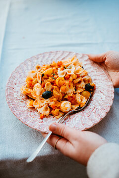 person holding pink plate with mixed pasta and black olives.