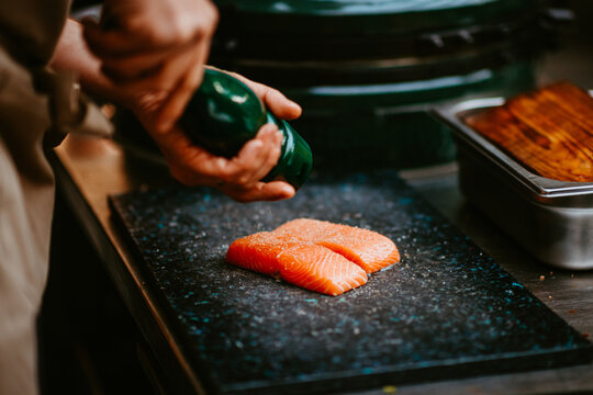 chef's hands seasoning salmon fillets with pepper by grill.